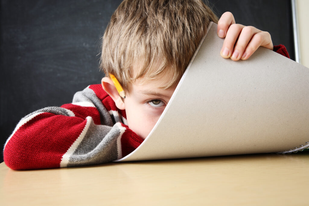 A frustrated child places his chin on the desk and curls up a pad of paper to partly obscure his face.
